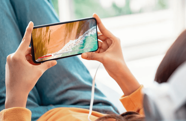 มือที่กำลังถือสมาร์ทโฟนในแนวนอนเพื่อรับชมวิดีโอชายหาดและคลื่นทะเล แสดงให้เห็นถึงประสบการณ์หน้าจอและการใช้งานที่สะดวกสบาย ซึ่งเป็นประเด็นสำคัญในการเปรียบเทียบ vivo Y29 vs Y28