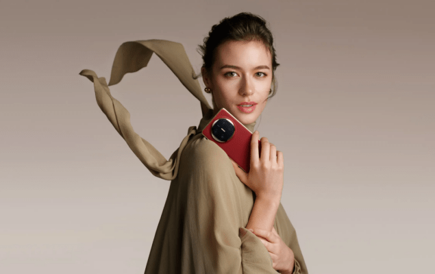 HUAWEI vs iPhone - ภาพผู้หญิงสวมเสื้อผ้าโทนสีอ่อนและมีผ้าพันคอปลิวไหว กำลังถือสมาร์ทโฟน HUAWEI สีแดงโดดเด่นที่มีโมดูลกล้องทรงกลม/วงแหวนอยู่บริเวณไหล่ ซึ่งแสดงถึงดีไซน์ที่หรูหราและเน้นกล้องเพื่อเปรียบเทียบในบทความ HUAWEI vs iPhone
