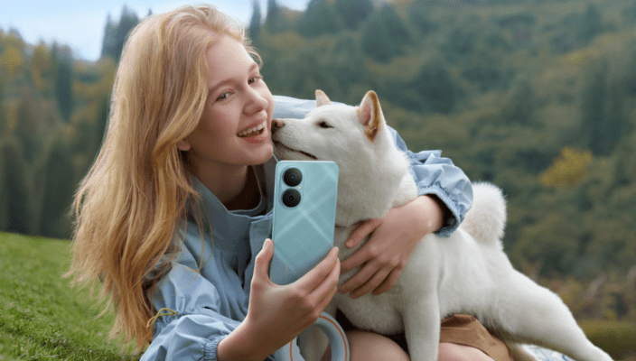 ผู้หญิงผมบลอนด์กำลังถ่ายเซลฟี่กับสุนัขสีขาว โดยถือสมาร์ทโฟน HUAWEI สีฟ้าอ่อน ซึ่งเป็นภาพที่แสดงถึงการใช้งานโทรศัพท์ในชีวิตประจำวันเพื่อตอบคำถามโทรศัพท์ HUAWEI ดีไหม