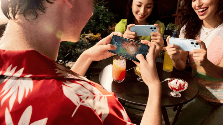 ภาพคนกลุ่มหนึ่งกำลังใช้สมาร์ทโฟนถ่ายรูปและเล่นเกมกลางแจ้ง สื่อถึงประสิทธิภาพด้านความบันเทิงและการใช้งานแบตเตอรี่ของ Samsung Galaxy A56 5G vs OPPO Reno14 F 5G