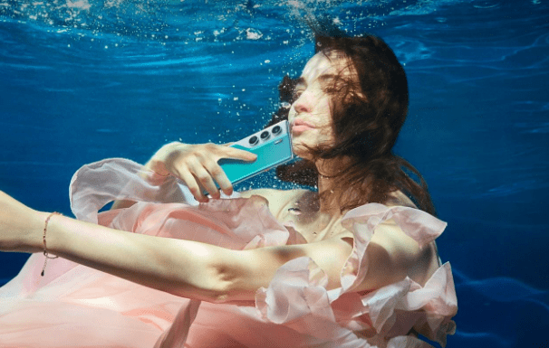 รีวิว TECNO Camon 40 Pro – ผู้หญิงในชุดสีชมพูกำลังถ่ายภาพตัวเองใต้น้ำโดยถือโทรศัพท์มือถือ TECNO Camon 40 Pro สีฟ้าหรือเขียว เพื่อเน้นความสามารถในการกันน้ำและประสิทธิภาพกล้องสำหรับการถ่ายภาพในสภาพแวดล้อมที่ท้าทาย