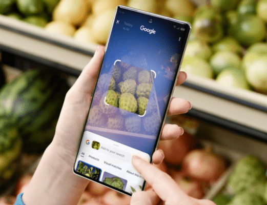 รีวิว TECNO Camon 40 Pro – มือที่กำลังถือโทรศัพท์มือถือ TECNO Camon 40 Pro เพื่อใช้แอปพลิเคชัน Google Lens สแกนผลไม้ในซูเปอร์มาร์เก็ต เพื่อเน้นฟีเจอร์กล้องอัจฉริยะและการประมวลผลของเครื่อง