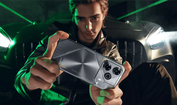 นายแบบหนุ่มสไตล์ดุดันกำลังชูโทรศัพท์มือถือ TECNO POVA 6 Pro 5G สีเทาเข้ม ซึ่งมีลวดลายแบบเกมมิ่ง/ไซเบอร์พันธงที่ฝาหลังอย่างชัดเจน ภายใต้แสงไฟสปอร์ตไลท์สีเขียวนีออน และฉากหลังเป็นกระจังหน้ารถสปอร์ต รูปภาพนี้เน้นประสิทธิภาพสำหรับเล่นเกม ซึ่งเป็นปัจจัยหนึ่งที่ต้องพิจารณาเมื่อสงสัยว่า โทรศัพท์ TECNO ดีไหม