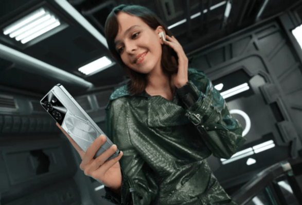รีวิว TECNO Pova 5 Pro 5G – ผู้หญิงกำลังยิ้มและฟังเพลงด้วยหูฟังไร้สาย พร้อมถือโทรศัพท์มือถือ TECNO Pova 5 Pro 5G สีเงิน/เทา ที่แสดงด้านหลังดีไซน์ Mecha และไฟ LED ในสภาพแวดล้อมสไตล์ไซไฟ