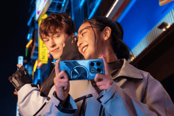 ชายหญิงสองคนกำลังถ่ายรูปหรือดูโทรศัพท์มือถือ Infinix ที่มีดีไซน์ฝาหลังแบบ Cyber-Mecha และไฟวงกลมที่โดดเด่นในเวลากลางคืนที่มีแสงไฟนีออน ภาพนี้แสดงถึงการใช้งานสมาร์ทโฟนของ Infinix ในสภาพแสงน้อย ซึ่งเป็นปัจจัยสำคัญในการเปรียบเทียบในบทความ TECNO vs Infinix
